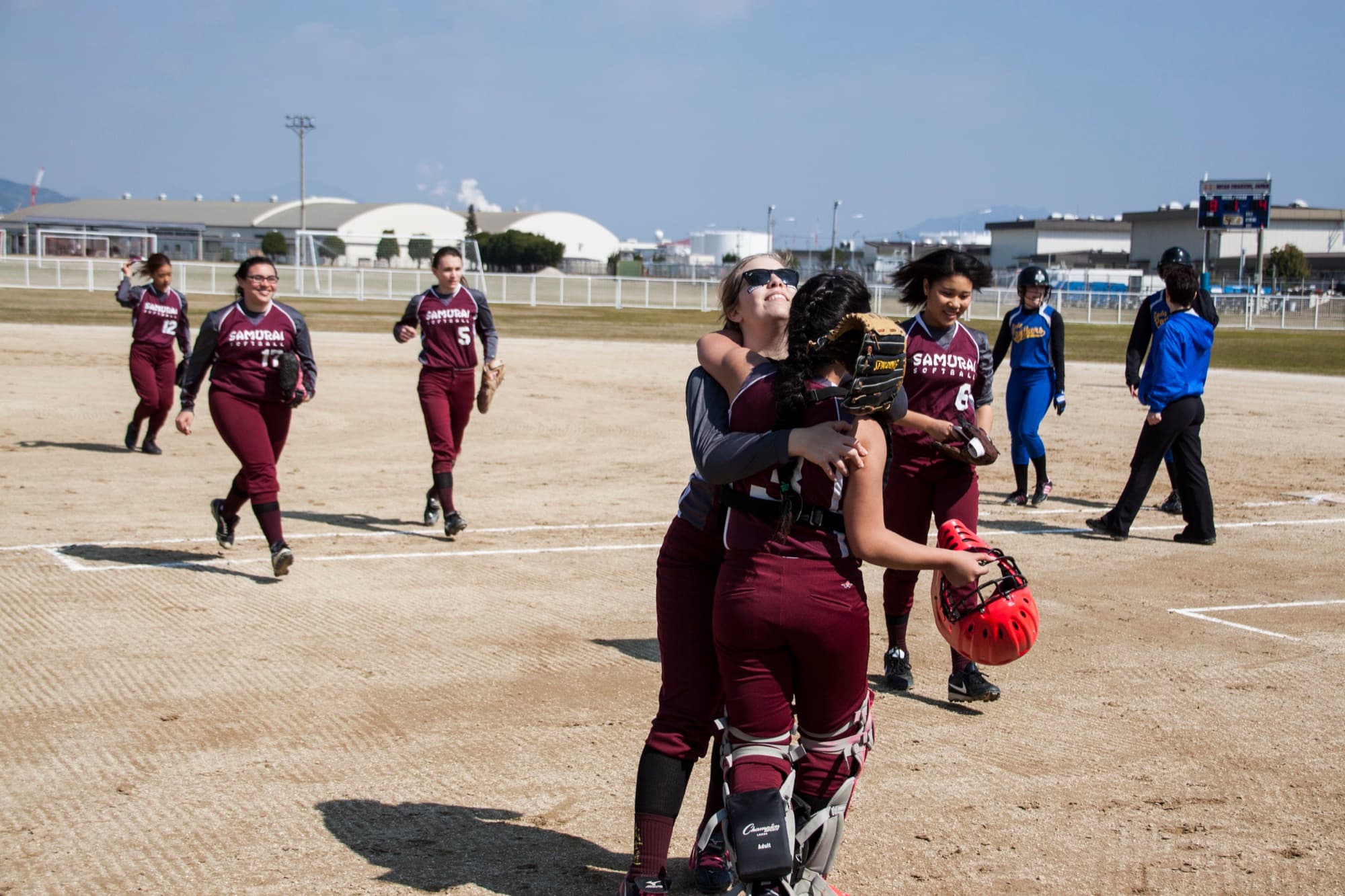 Two DoDEA high school softball teammates in civilian sports uniforms hugging after a game