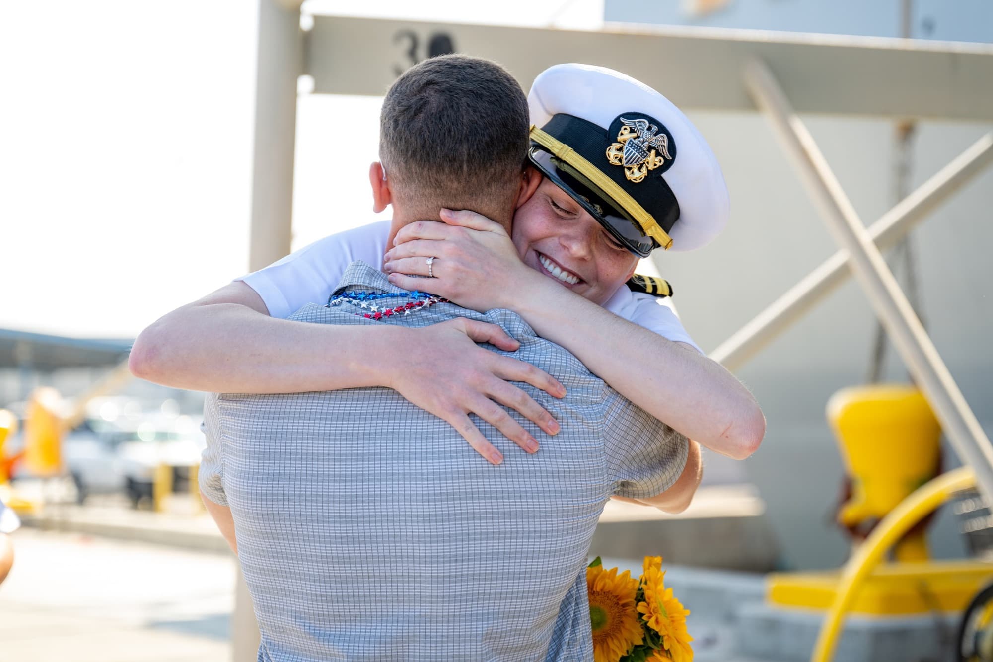 A Navy officer in dress whites embraces her civilian husband on the pier after a homecoming